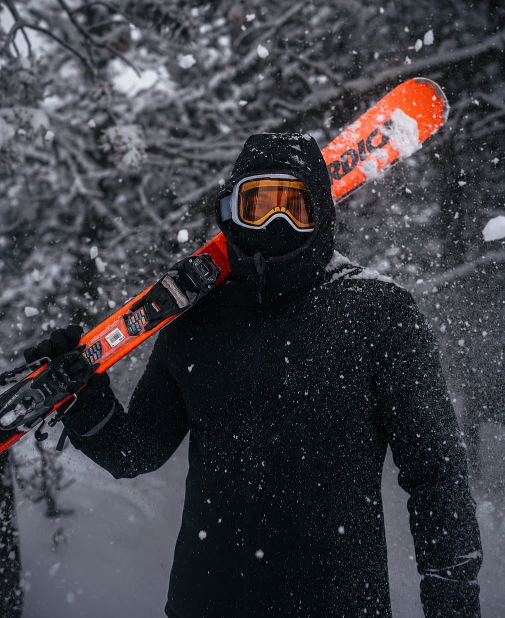 Skieur masqué en tenue noire portant des skis orange sur l’épaule sous une chute de neige